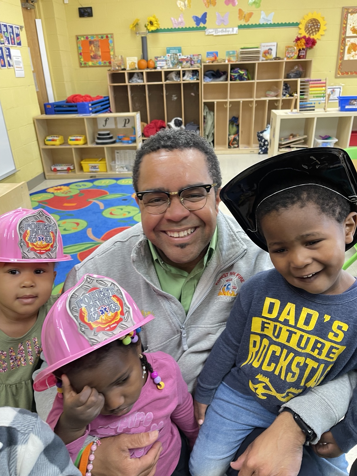 A smiling Forest Park firefighter with children wearing pink and black firefighter hats in a Learning Garden classroom