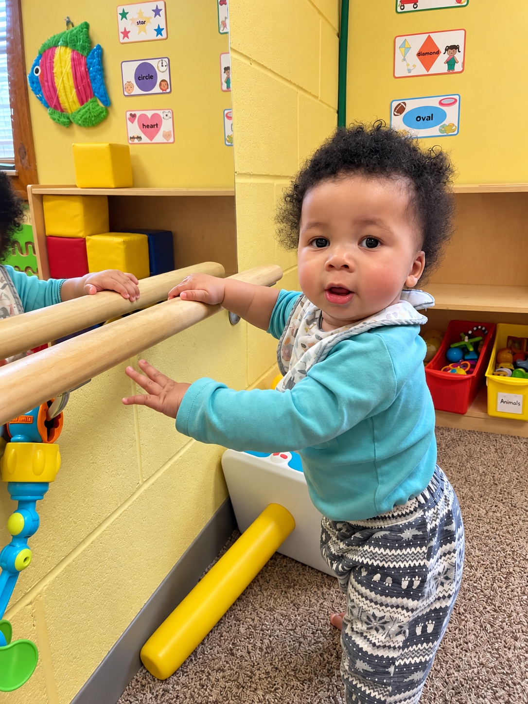 A baby practices pulling up to stand beside a classroom shelf.